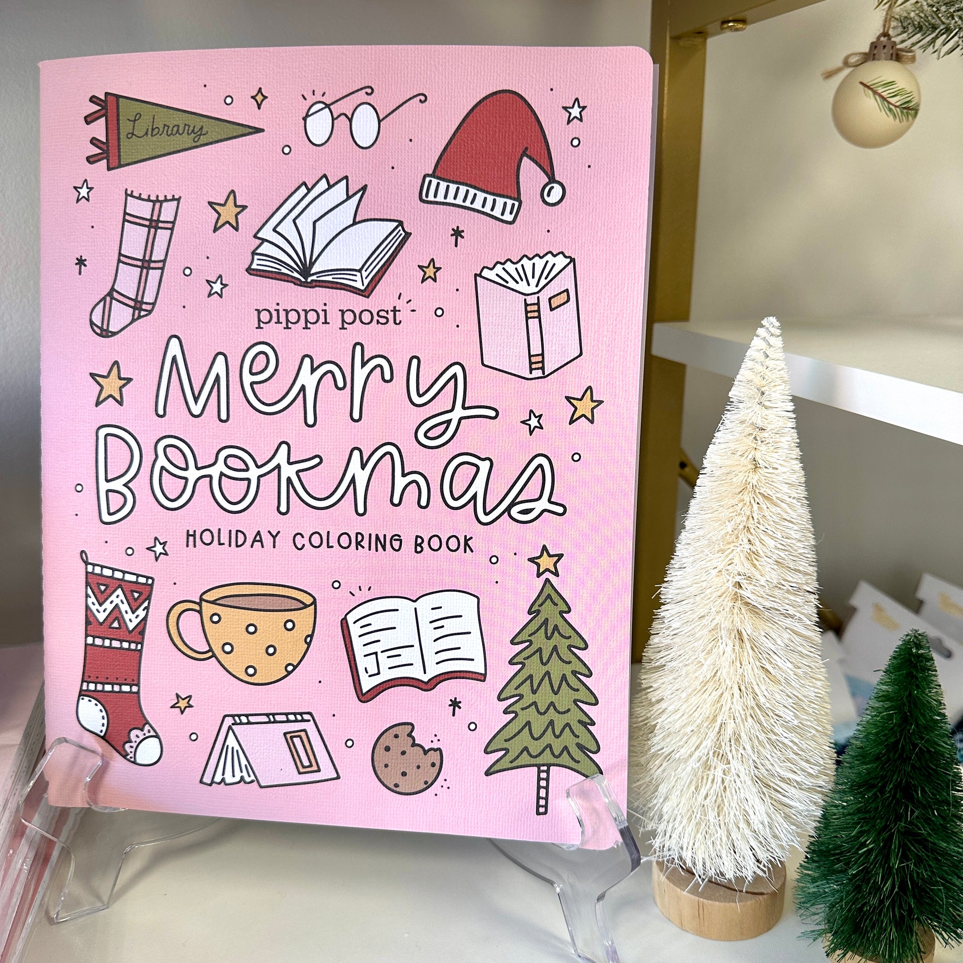 Pink 'Merry Bookmas' holiday coloring book with Christmas-themed illustrations on a stand.