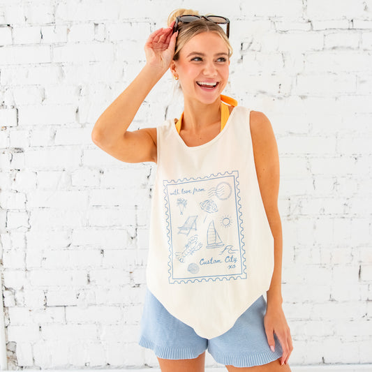 girl standing in front of a white brick wall. Her hair is pulled up into a messy bun with a clip, she has sunglasses resting on top of her head. She's wearing light blue knit shorts and an oversized tank top over an orange bikini. The tank top features a custom printed "With love from" tank top. The design on the tank top has a large stamp outline with fun coastal, beachy icons inside as well as a spot to add a custom city. The tank top is ivory and the print is blue.