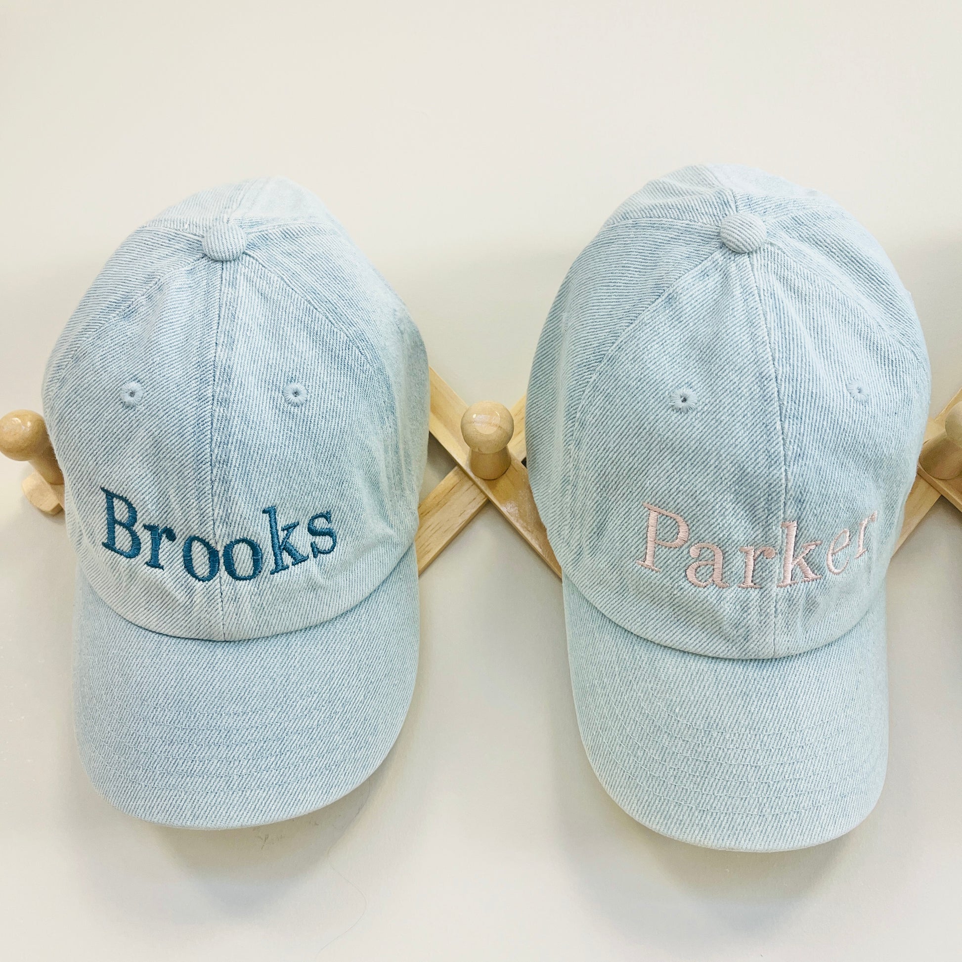 Two light blue baseball caps with embroidered names on a wooden stand.