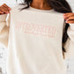 close up of a girl standing in front of brick wall wearing a sweet cream crewneck sweatshirt with INTROVERTED (but willing to discuss books) embroidered across the chest in a powder pink thread