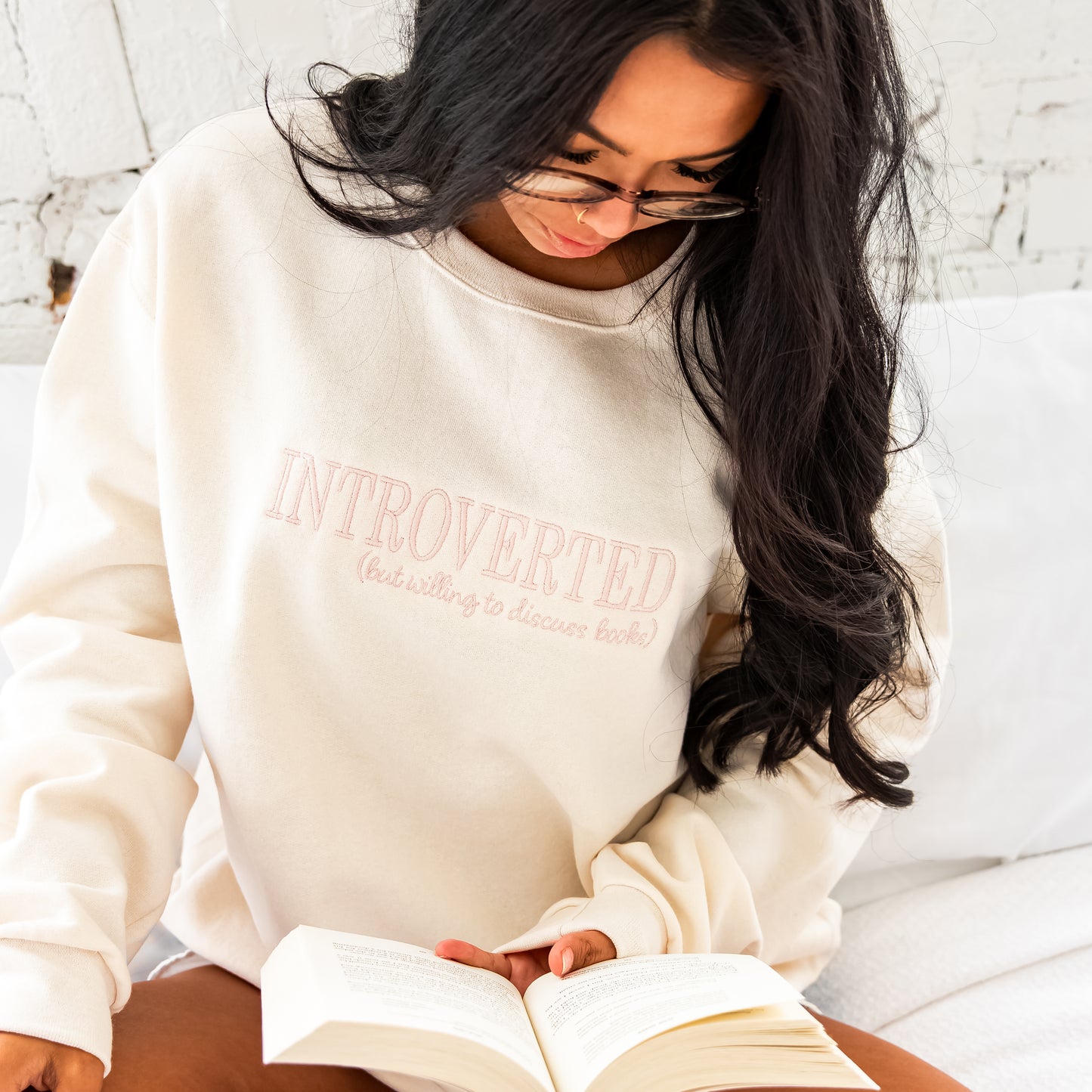 girl sitting on a bed in front of brick wall, reading a book, wearing a sweet cream crewneck sweatshirt with INTROVERTED (but willing to discuss books) embroidered across the chest in a powder pink thread