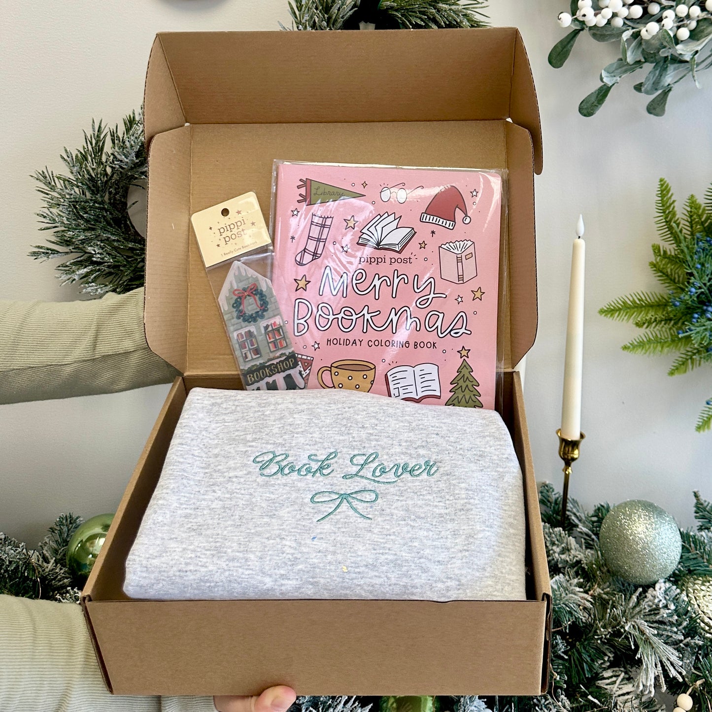 Box containing a 'Book Lover' sweatshirt and 'Merry Bookmas' coloring book and bookshop bookmark with Christmas decorations in the background.