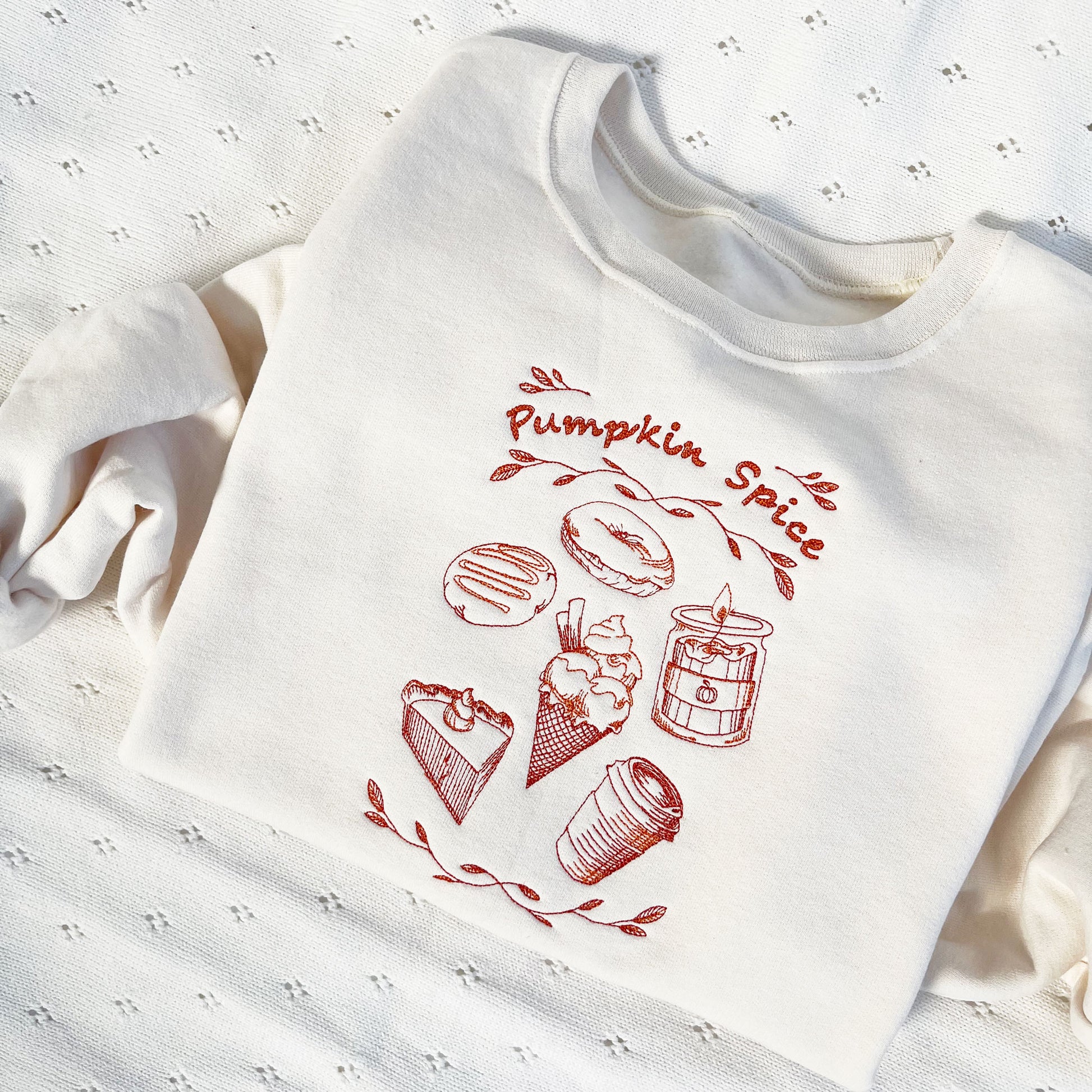 A sweet cream crewneck sweatshirt with a autumnal stitched pumpkin spice design featuring a donut, candle, ice cream, pie, and a coffee cup in a vibrant pumpkin spice and rust colored thread.