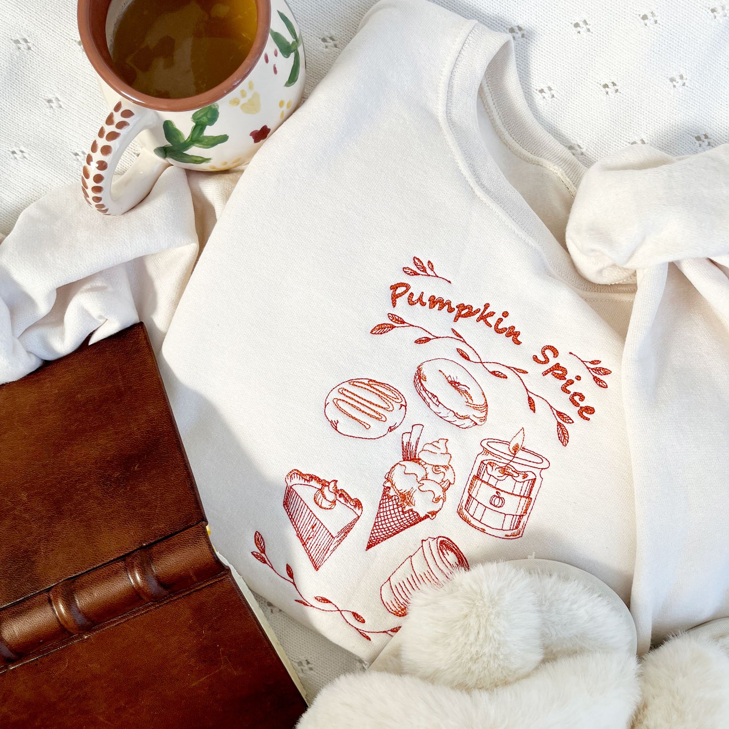 flat lay featuring a leather book, coffee, slippers, and a sweet cream crewneck sweatshirt with a autumnal stitched pumpkin spice design featuring a donut, candle, ice cream, pie, and a coffee cup