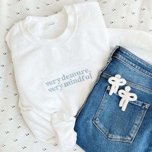 outfit flat lay featuring jeans, white bow earrings, and a white crewneck sweatshirt with very demure, very mindful embroidered in a powder blue thread.