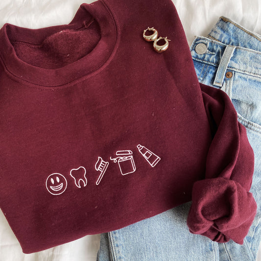 maroon crewneck sweatshirt with embroidered mini dental icons across the chest in white thread styled with blue jeans and gold hoop earings