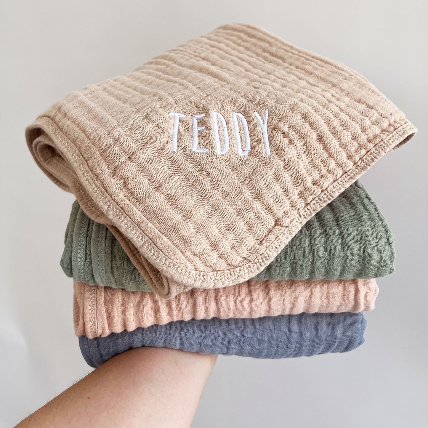 stack of muslin baby blankets in blue, pink, green, and sand. Top sand blanket showing an embroidered name in handicraft font and white thread.