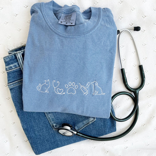 flat lay of a washed denim tee with embroidered veterinarian icons across the chest in white thread.