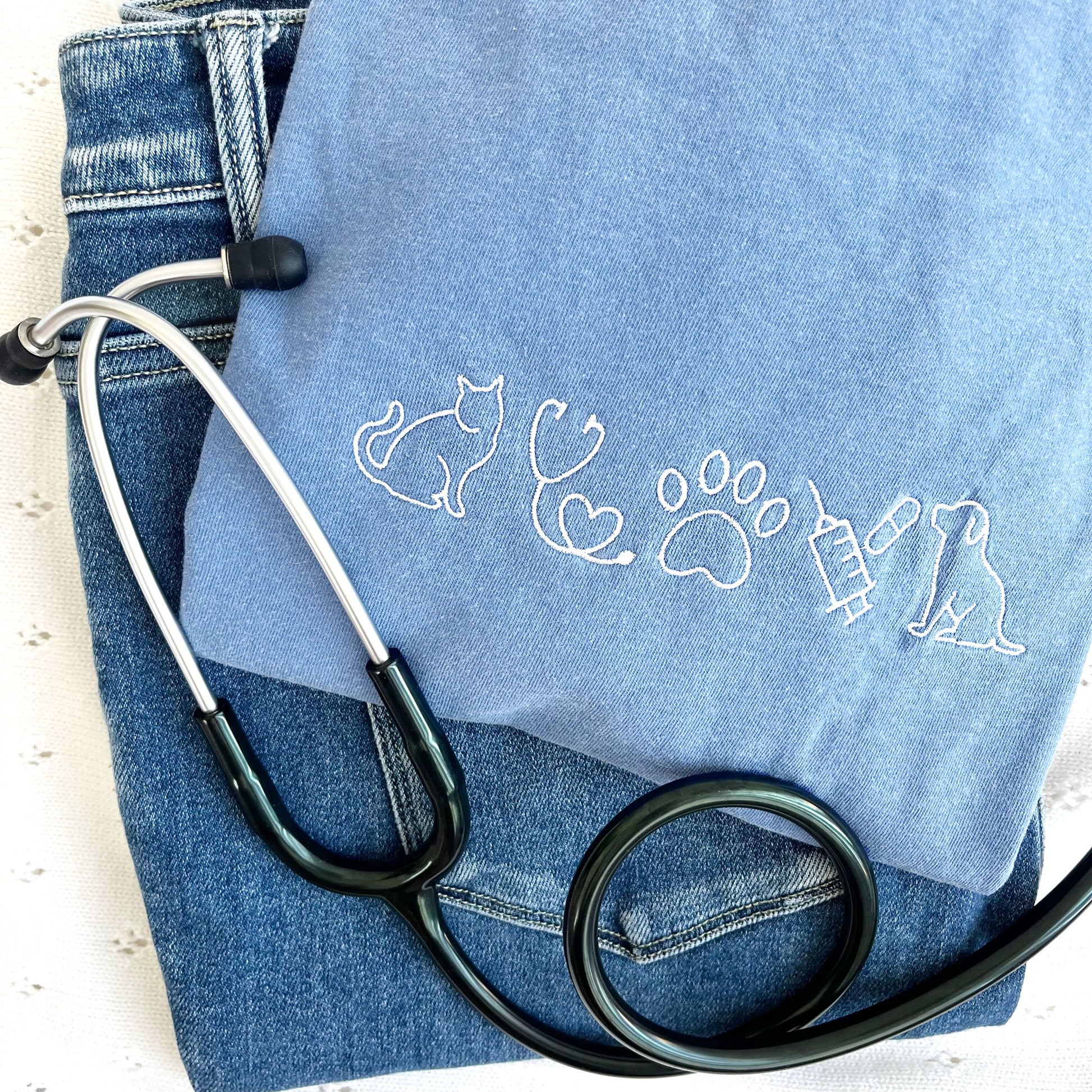 flat lay of a washed denim tee with embroidered veterinarian icons across the chest in white thread.