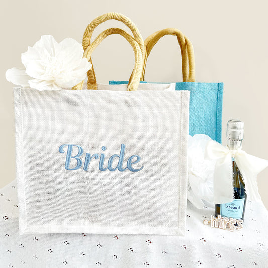 White and Palace Blue jute gift tote with a custom bride embroidery, paired with a bottle of champagne and a pair of earrings