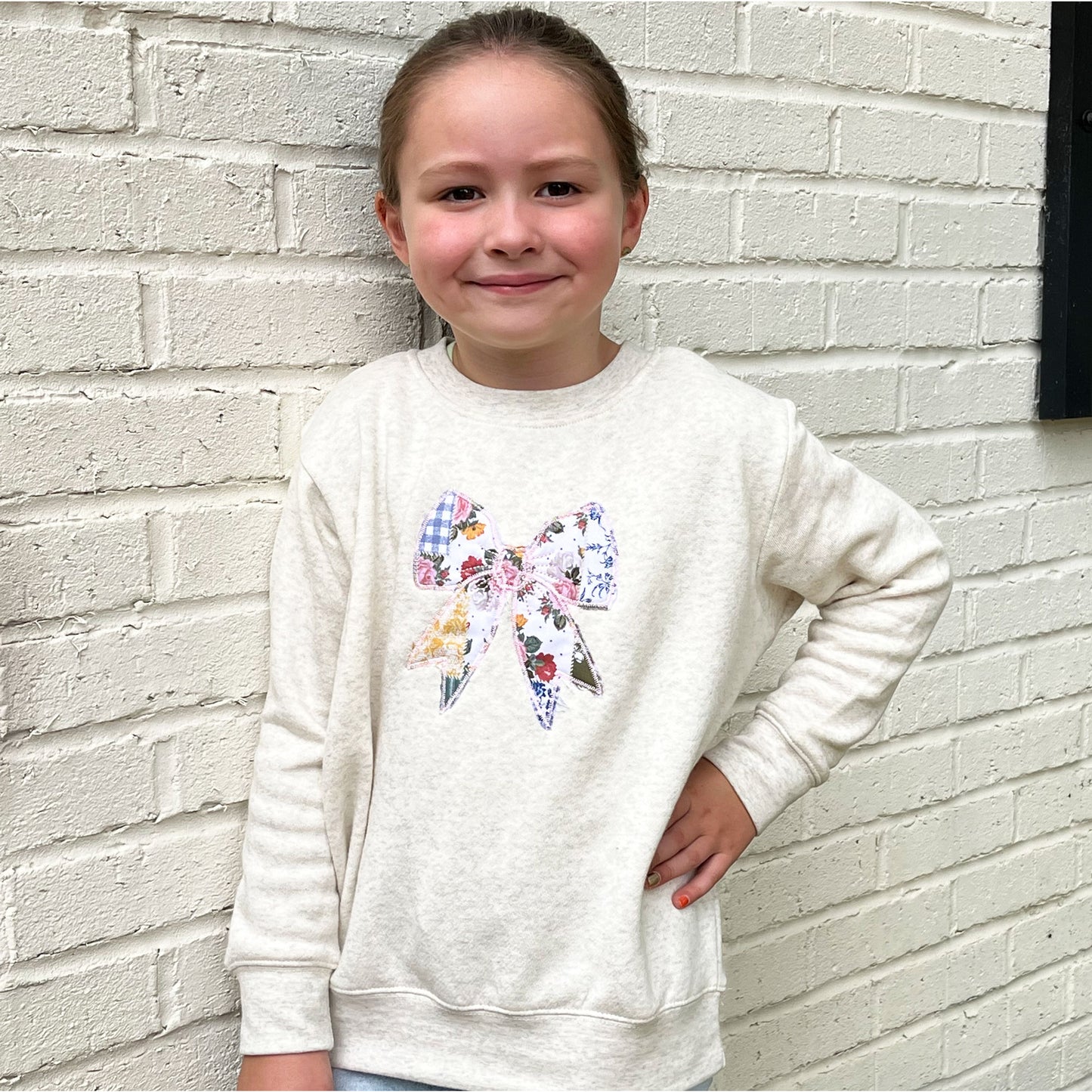 little girl wearing an oatmeal heather crewneck with custom embroidered patchwork quilted bow embroidered design