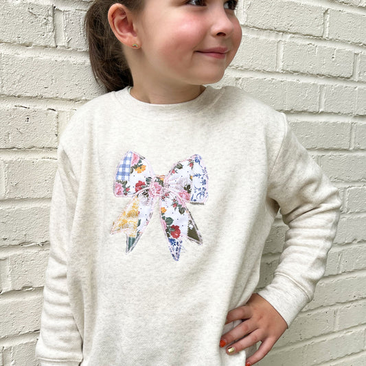 little girl wearing an oatmeal heather crewneck with custom embroidered patchwork quilted bow embroidered design