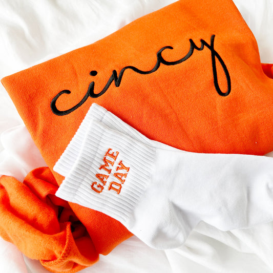 orange crewneck sweatshirt with cincy embroidered in black paired with custom embroidered game day ankle crew socks