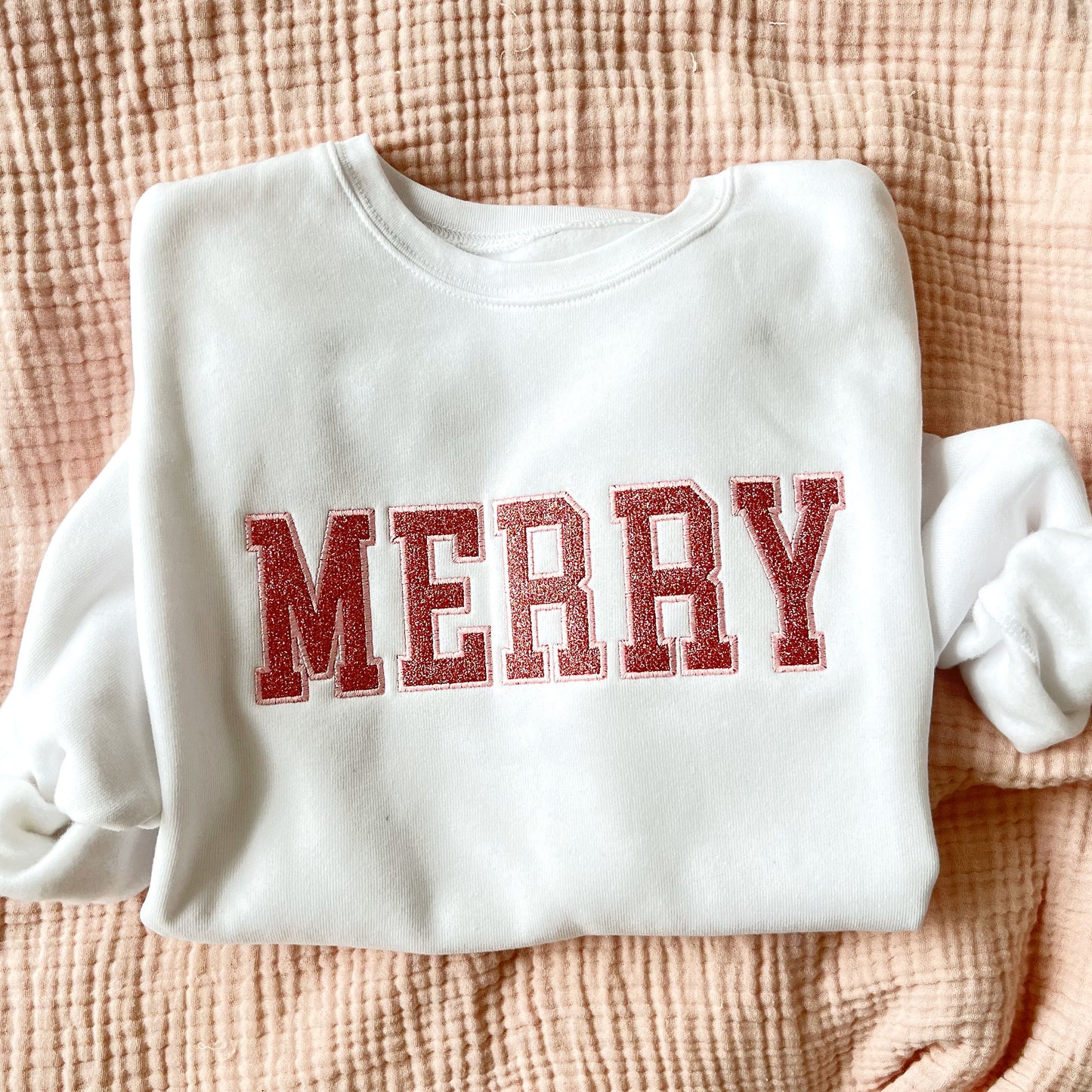 flat lay of a white crewneck sweatshirt with embroidered merry all caps in outline block font. Filled in the text is a pink glitter fill.