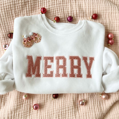 flat lay of a white crewneck sweatshirt with embroidered merry all caps in outline block font. Filled in the text is a pink glitter fill.