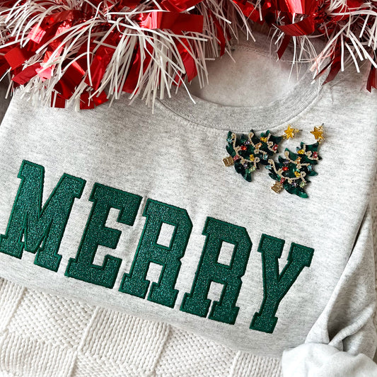 close up of a flat lay of an ash crewneck sweatshirt with embroidered merry in all caps and green thread. In the outline embroidered text is a glittery green fill. The sweatshirt is styled with Christmas tree earrings and red garland.