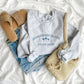 styled flat lay with jean shorts, a tote bag, sandals, and an ash crewneck sweatshirt. the sweatshirt has honeymoon island state park and alm tre design embroidered in french blue thread across  the chest