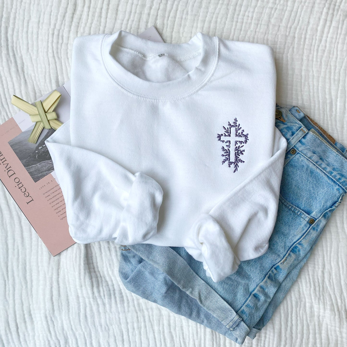flat lay of a prayer book, palm cross, jean shorts, and a white crewneck sweatshirt with a floral cross embroidered on the left chest in smoky orchid thread