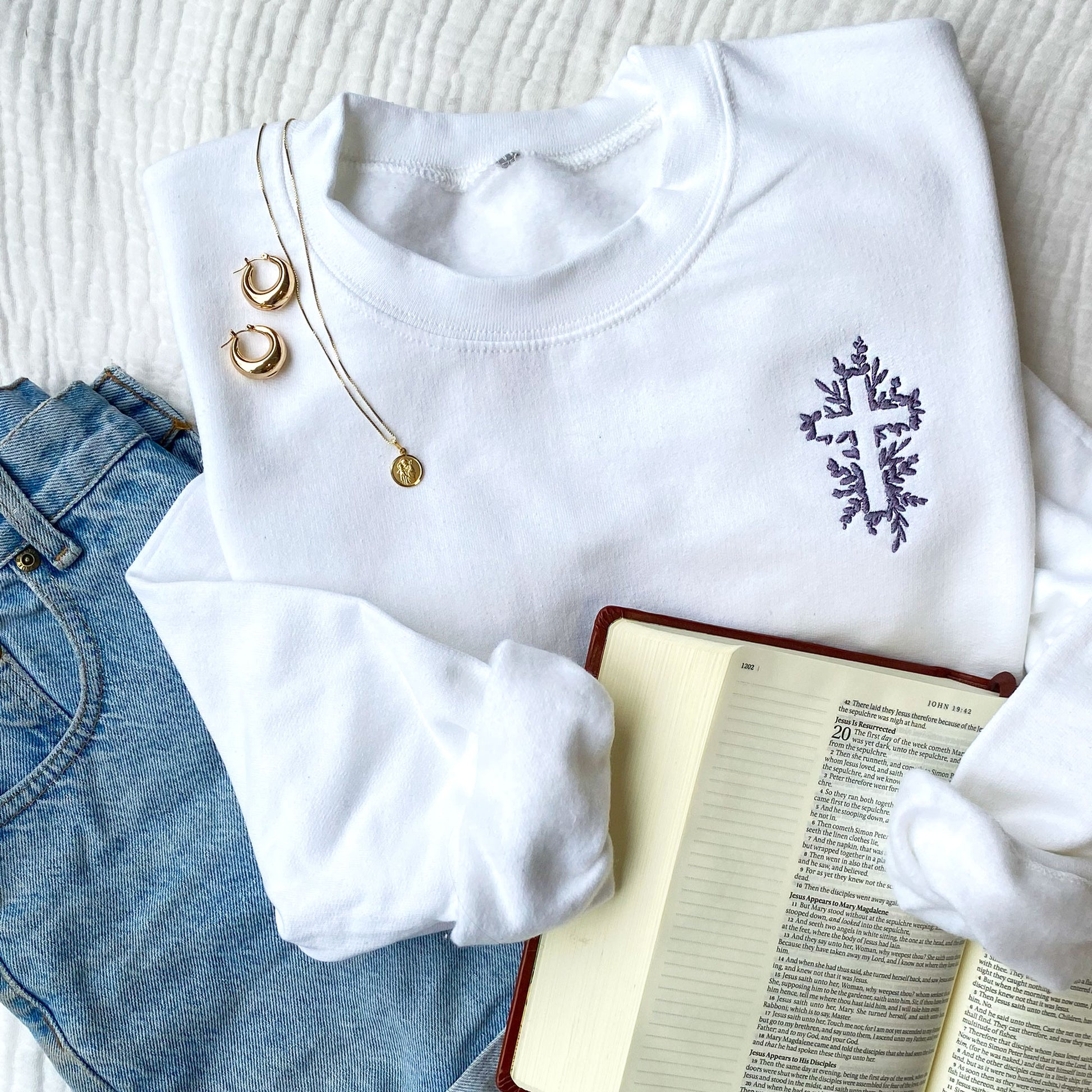 Flat lay of a white crewneck sweatshirt with a floral cross embroidered on the left chest in smoky orchid thread with a bible, jeans, and jewelry 