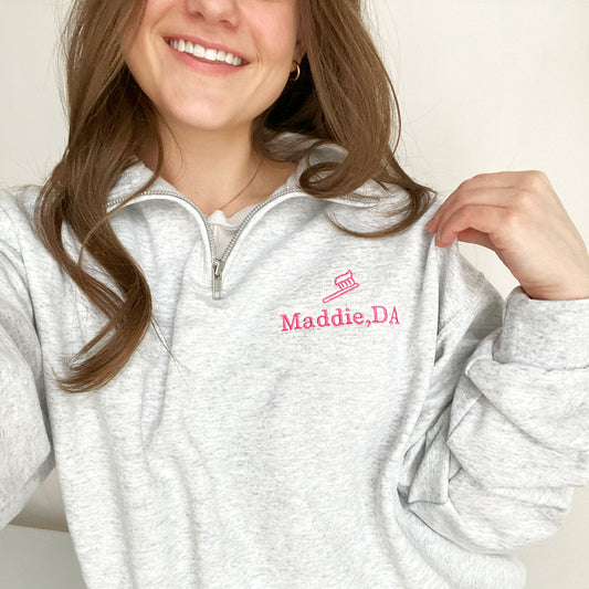 young woman wearing an ash quarterzip sweatshirt with pink embroidered tooth brush and name Maddie, DA on the left chest 