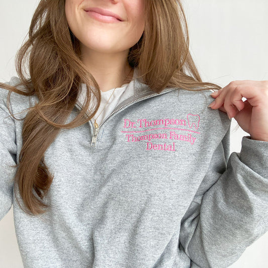 young woman wearing an oxford quarter zip sweatshirt. On the left chest in embroidered Dr. Thompson Thompson Family Dental and mini outline tooth in pink thread