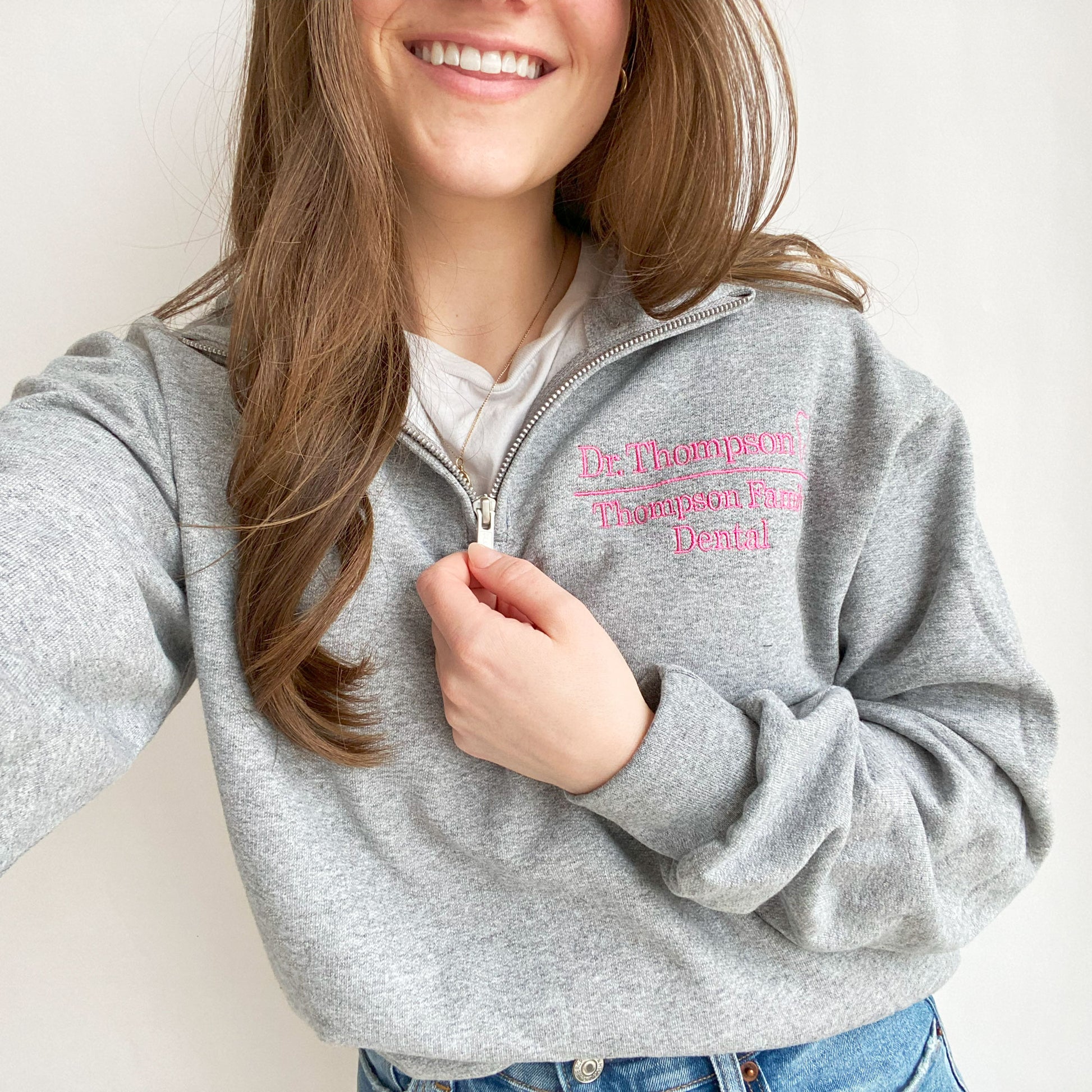young woman wearing an oxford quarter zip sweatshirt. On the left chest in embroidered Dr. Thompson Thompson Family Dental and mini outline tooth in pink thread
