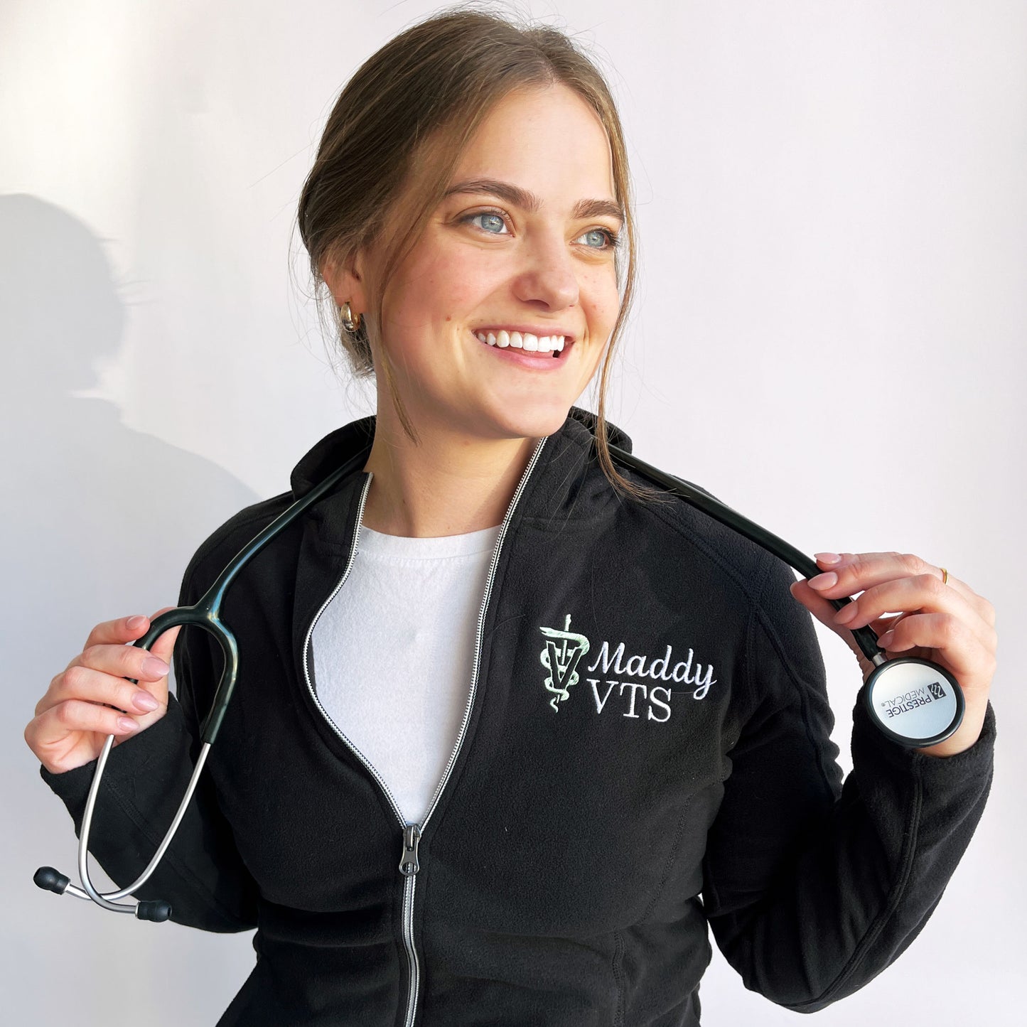 woman wearing a black full zip fleece jacket with custom name and veterinary medicine credentials and the veterinary caduceus symbol embroidered on the left chest in white and silver sage thread