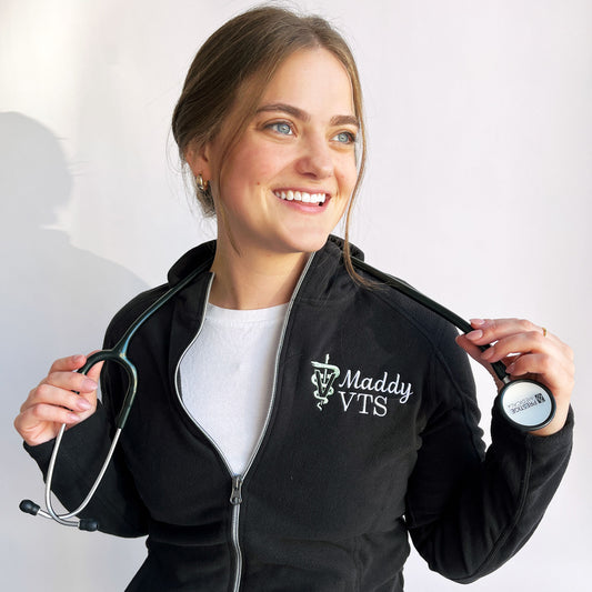 woman wearing a black full zip fleece jacket with custom name and veterinary medicine credentials and the veterinary caduceus symbol embroidered on the left chest in white and silver sage thread