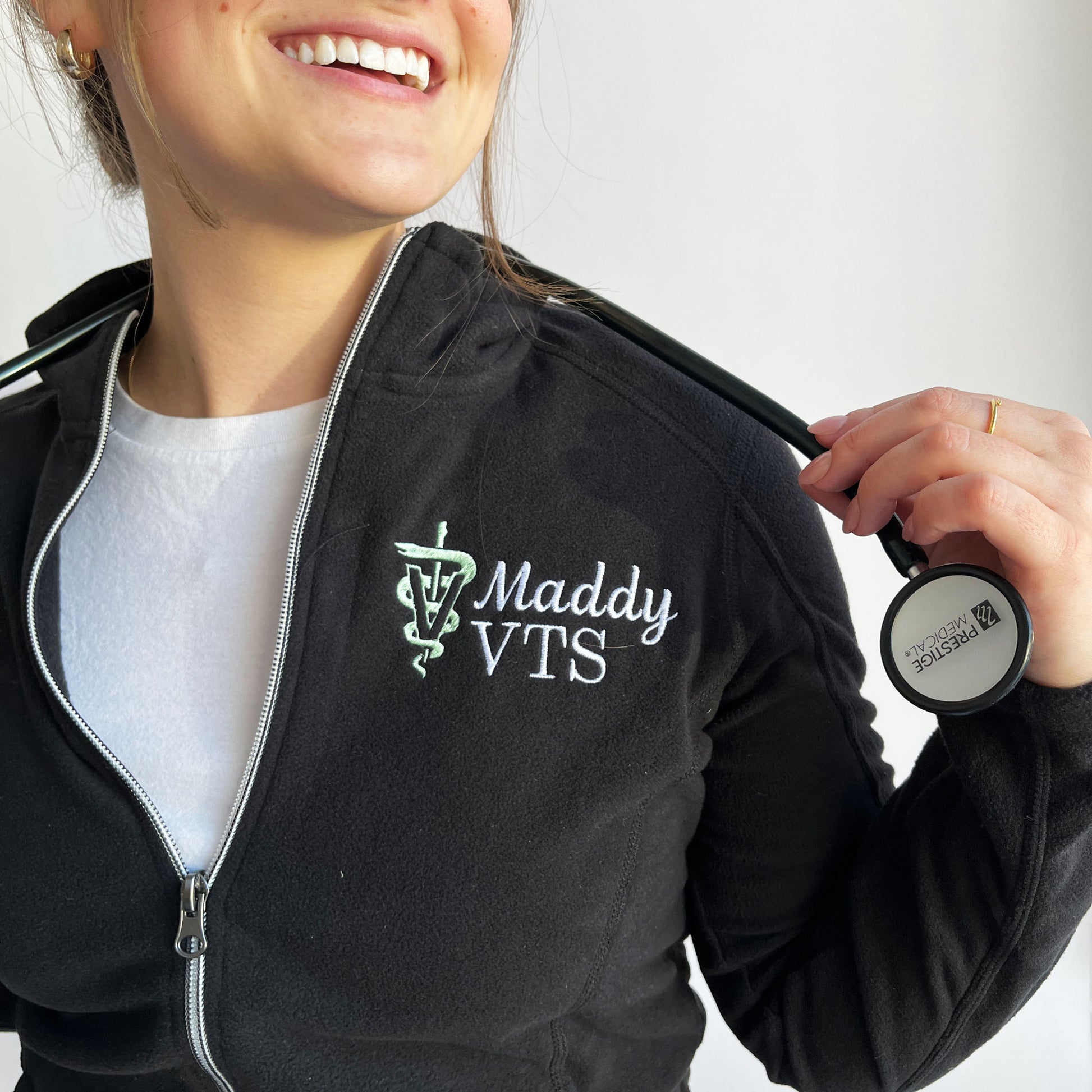 woman wearing a black full zip fleece jacket with custom name and veterinary medicine credentials and the veterinary caduceus symbol embroidered on the left chest in white and silver sage thread