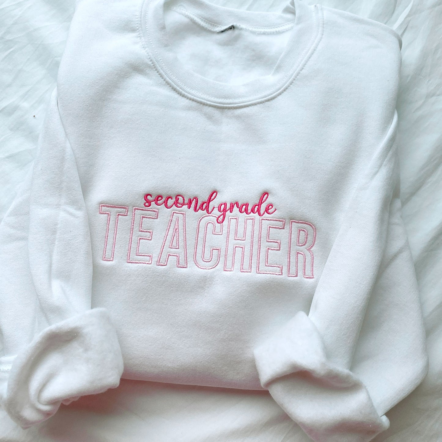 flat lay of a white crewneck sweatshirt with custom grade level and teacher embroidered design in pink and baby pink threads
