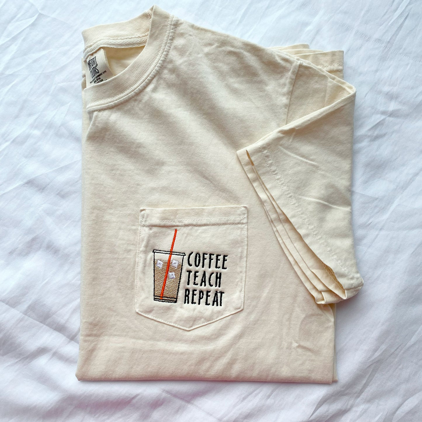 flat lay photo of an ivory comfort colors short sleeve t-shirt with an iced coffee with an orange straw and and coffee teach repeat text embroidered on the pocket