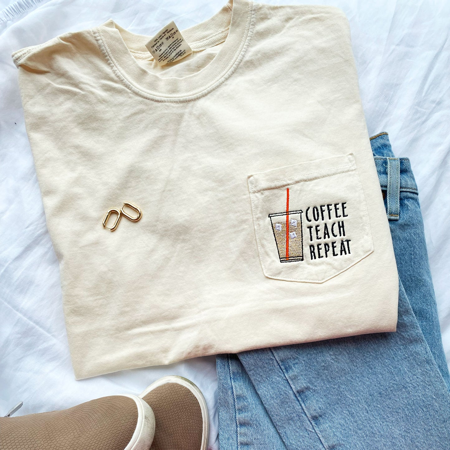 flat lay photo of an ivory comfort colors short sleeve t-shirt with an iced coffee with an orange straw and and coffee teach repeat text embroidered on the pocket