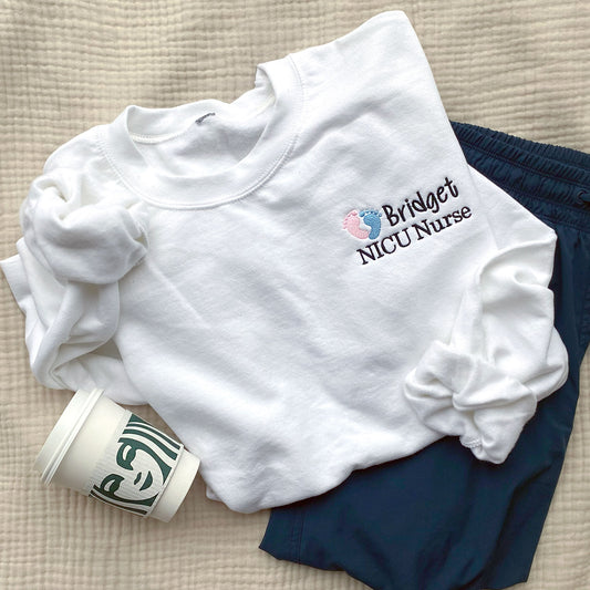 white crewneck sweatshirt styled with scrubs and a starbucks cup. embroidered on th e left chest is a mini nicu design with pink and blue baby feet and custom name and credentials in black thread 