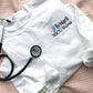 flat lay of a white crewneck sweatshirt with embroidered nicu design with pink and blue baby feet and custom name and credentials in black thread styled with a stethoscope