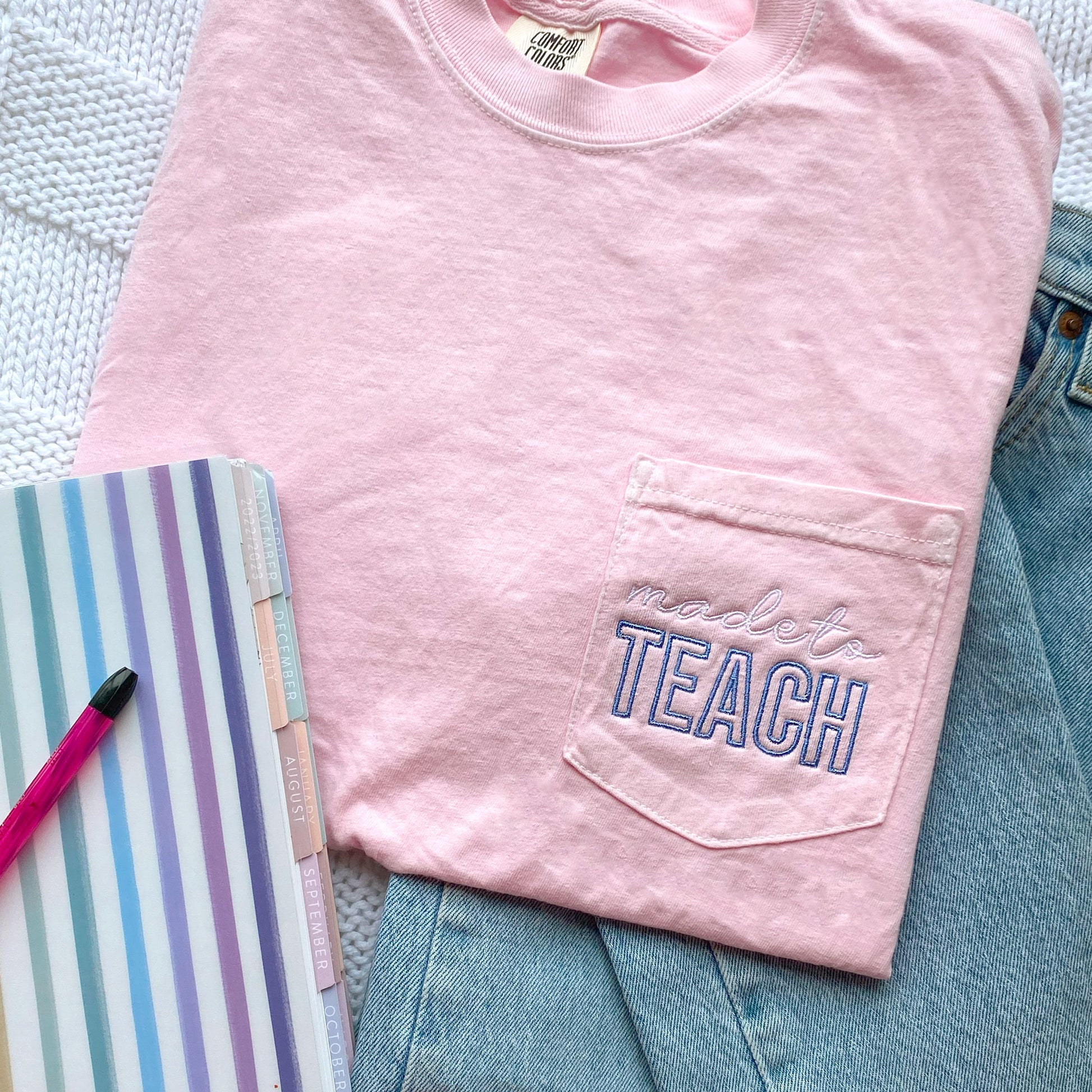 flat lay of a blossom comfort colors t-shirt with made to teach embroidered design on the pocket in white and periwinkle thread