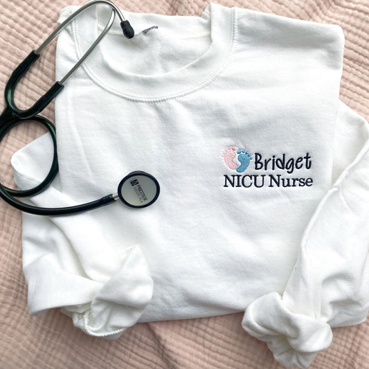 flat lay of a white crewneck sweatshirt  with embroidered nicu design with pink and blue baby feet and custom name and credentials in black thread styled with a stethoscope