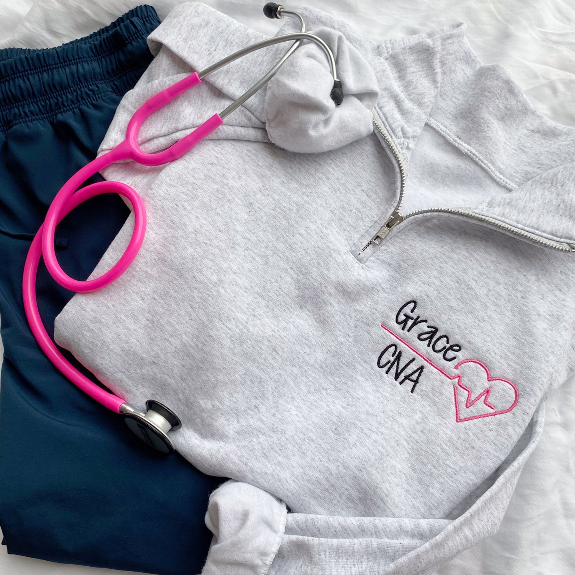 Ash quarterzip sweatshirt with custom nurse heart and heartbeat design in pink and name in credentials in black thread styled with scrubs and a stethoscope