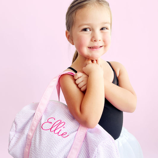 Little girl wearing a ballet outfit and holding a mini pink seersucker duffle bag with a custom name embroidered in a cute script font and pink thread.