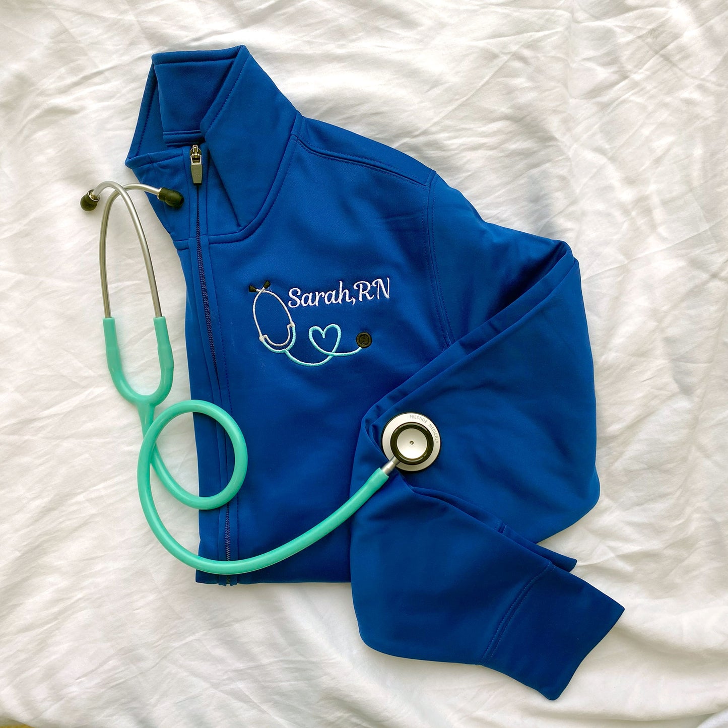 a royal blue full zip polyester nurse jacket with custom embroidered heart stethoscope and name in white and sky thread styled with a stethoscope.