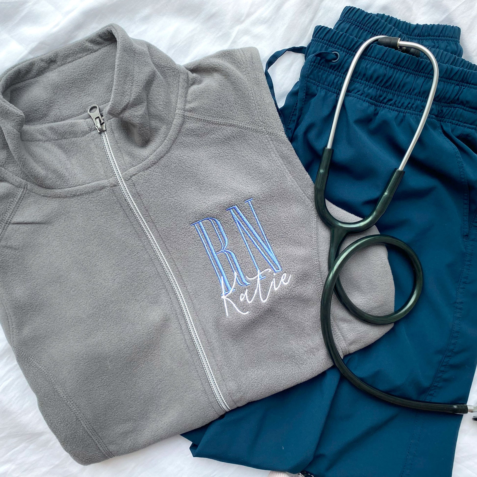 flat lay of  a grey fleece nurse vest with personalized RN design on the left chest in periwinkle and white with navy scrubs and a stethoscope
