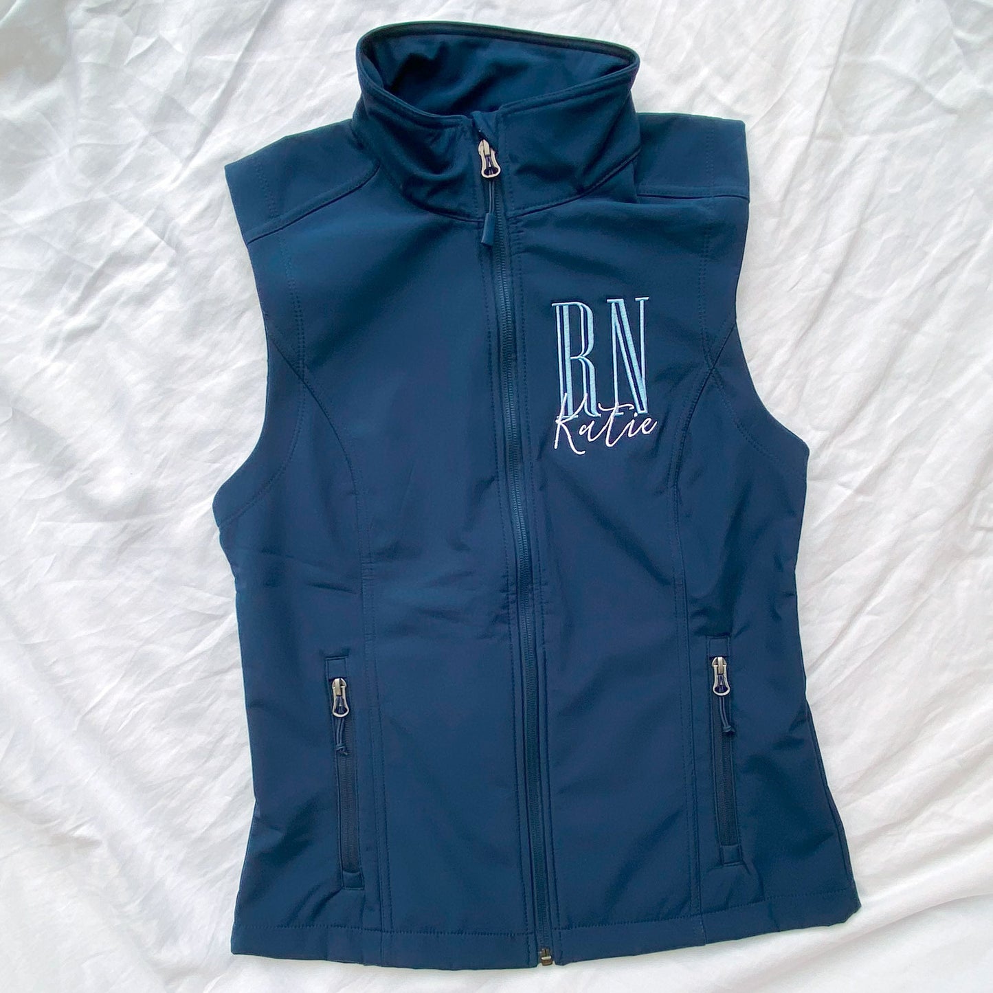 flat lay of a nurse navy soft shell vest with personalized embroidered RN and Name design on the left chest in baby blue and white threads