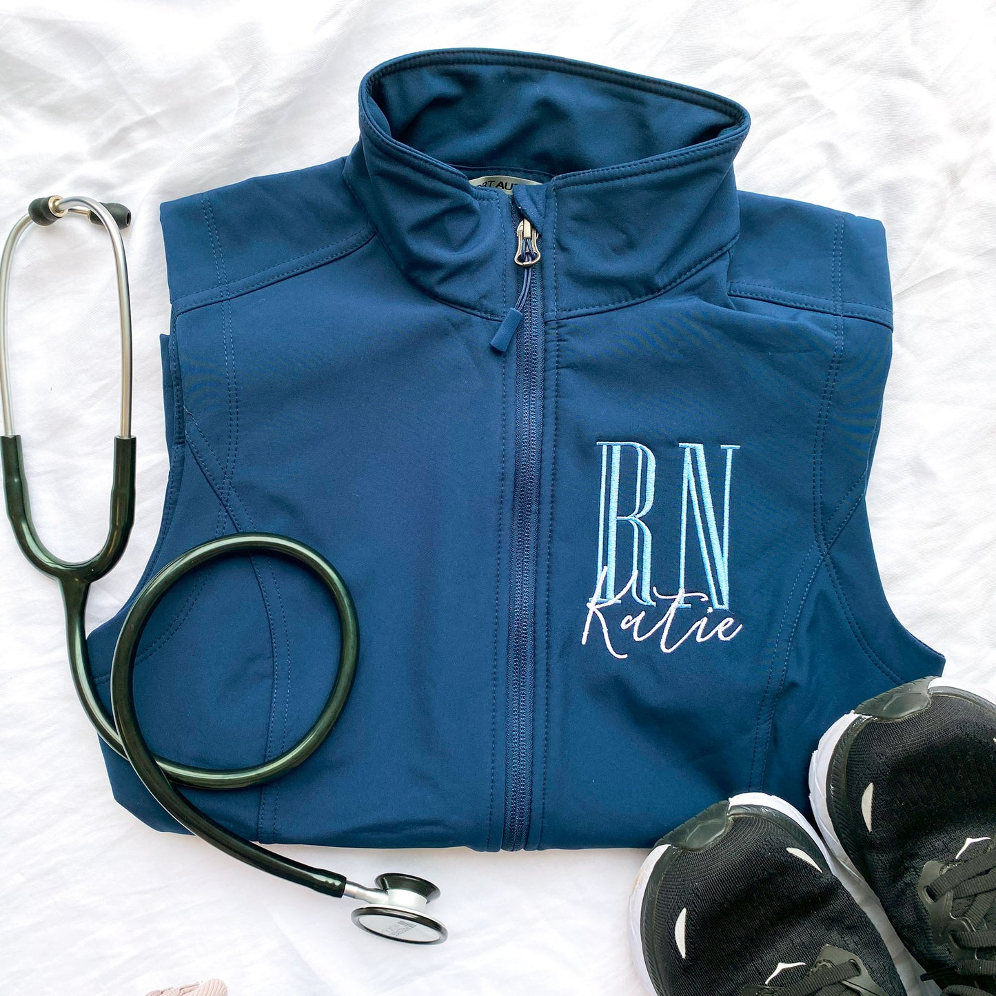 flat lay of a nurse navy soft shell vest with personalized embroidered RN and Name design on the left chest in baby blue and white threads 