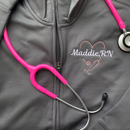 closeup of a grey polyester full zip with a cadet collar and a personalized heart shaped stethoscope and name embroidered design on the left chest.