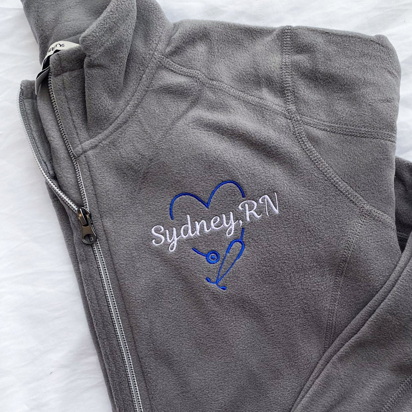 grey full zip fleece jacket with royal blue heart shaped stethoscope design with a custom name through the center embroidered on the left chest.