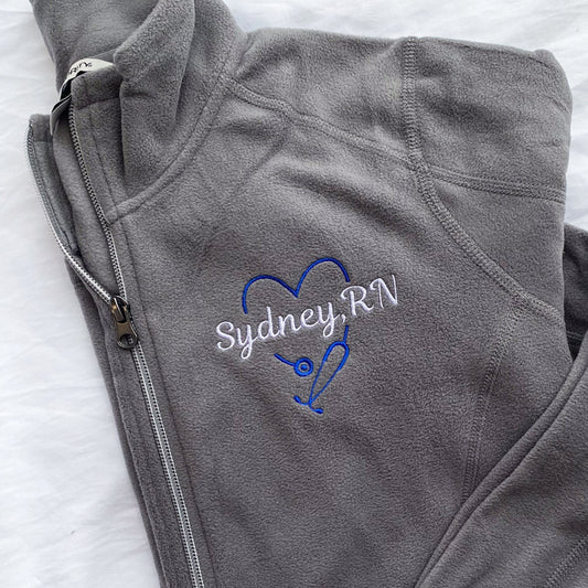 grey full zip fleece jacket with royal blue heart shaped stethoscope design with a custom name through the center embroidered on the left chest.