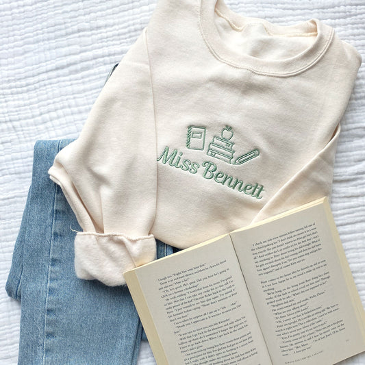 styled flat lay of a sweet cream crewneck sweatshirt with small embroidered teacher icons and custom name in silver sage thread across the chest with jeans and an open book