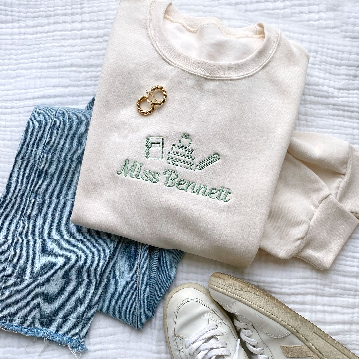 styled flat lay of a sweet cream crewneck sweatshirt with small embroidered teacher icons and custom name in silver sage thread across the chest with jeans and an open book