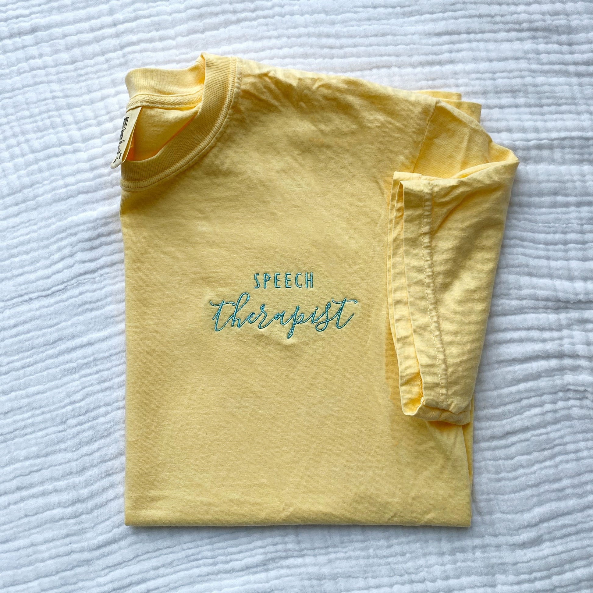flat lay of a custom embroidered speech therapist design in sky blue thread on a butter yellow comfort colors t-shirt