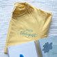 flat lay of a custom embroidered speech therapist design in sky blue thread on a butter yellow comfort colors t-shirt 