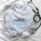flat lay of an ash crewneck sweatshirt with registered nurse embroidered design in black and baby blue threads